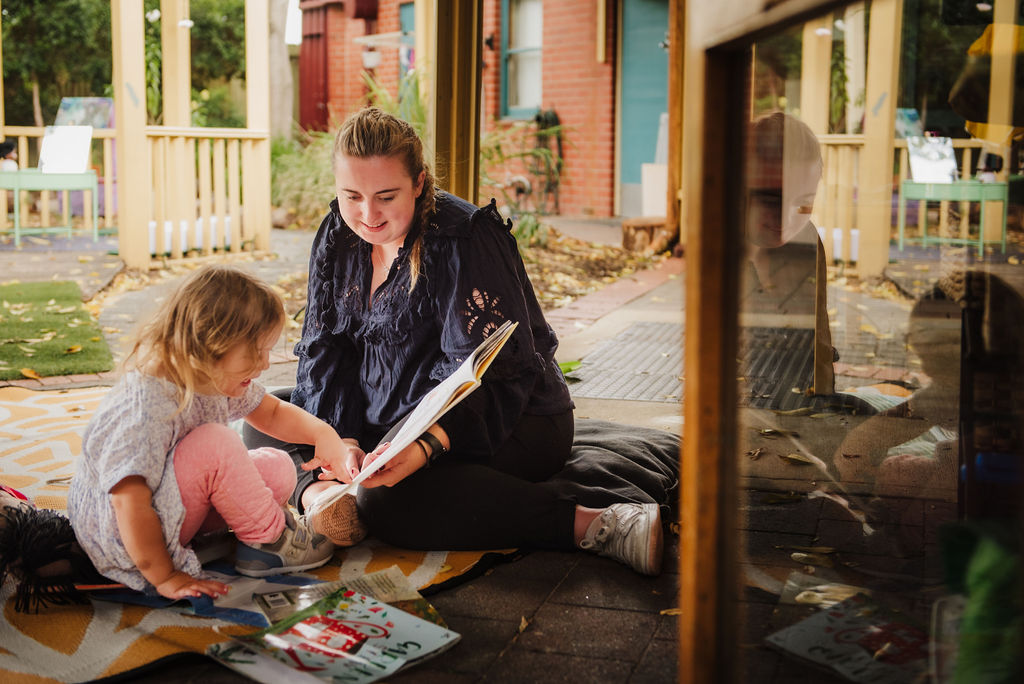 Child in a lilac dress and pink pants pointing at a book with an educator in a navy blue top