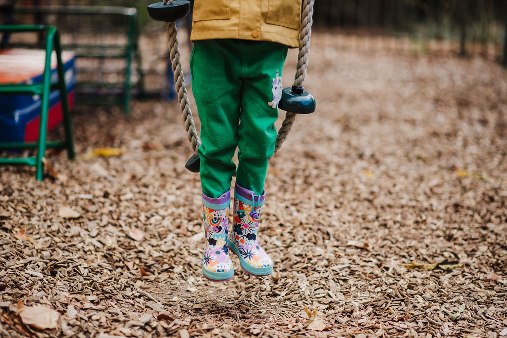 The legs of a child wearing bright green pants balancing on a rope bridge at a preschool playground