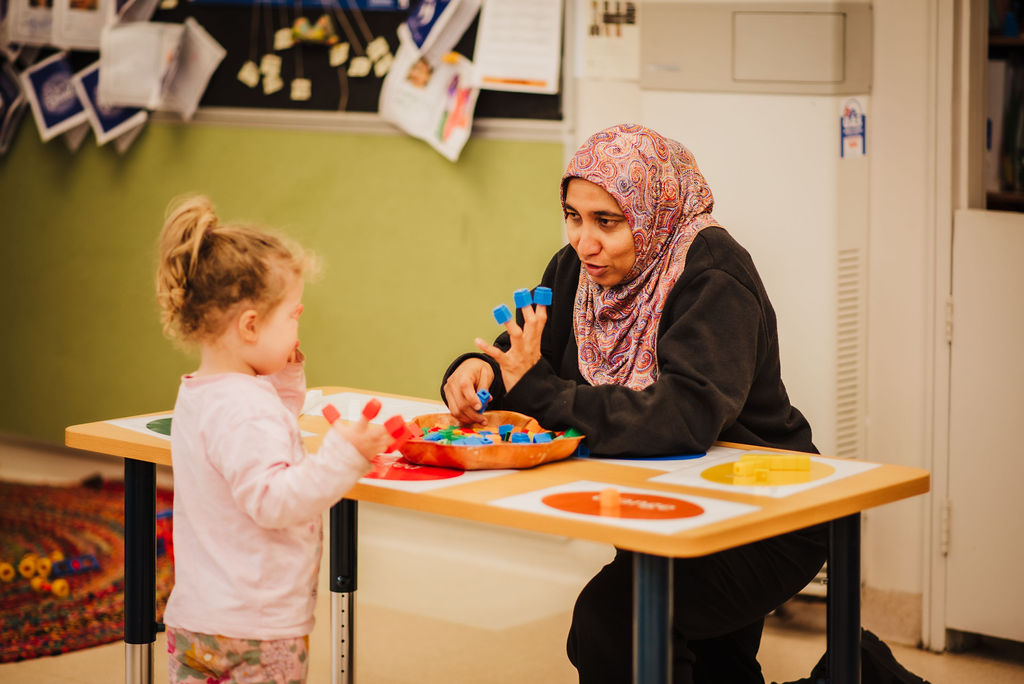 An educator wearing a pink patterned headscarf with blue blocks on her fingers with a small child with red blocks