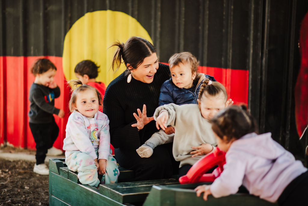 A young early childhood educator surrounded by toddlers with one in her arms