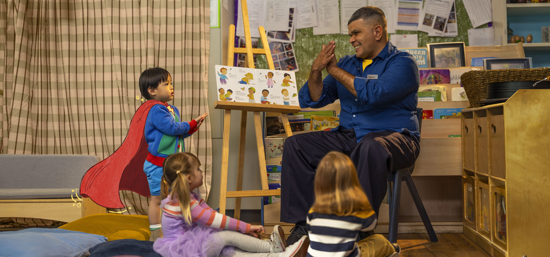 An Aboriginal man, an early childhood educator, reading a book to three children, who are sitting on the floor.