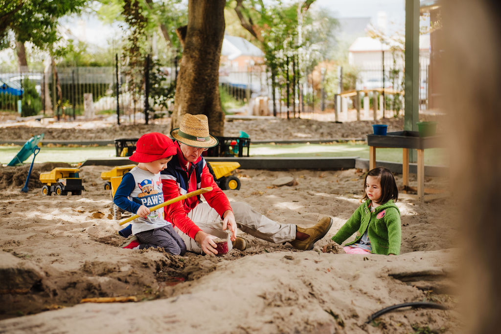Two children in the sandpit with an educator, playing with a yellow shovel in the sand